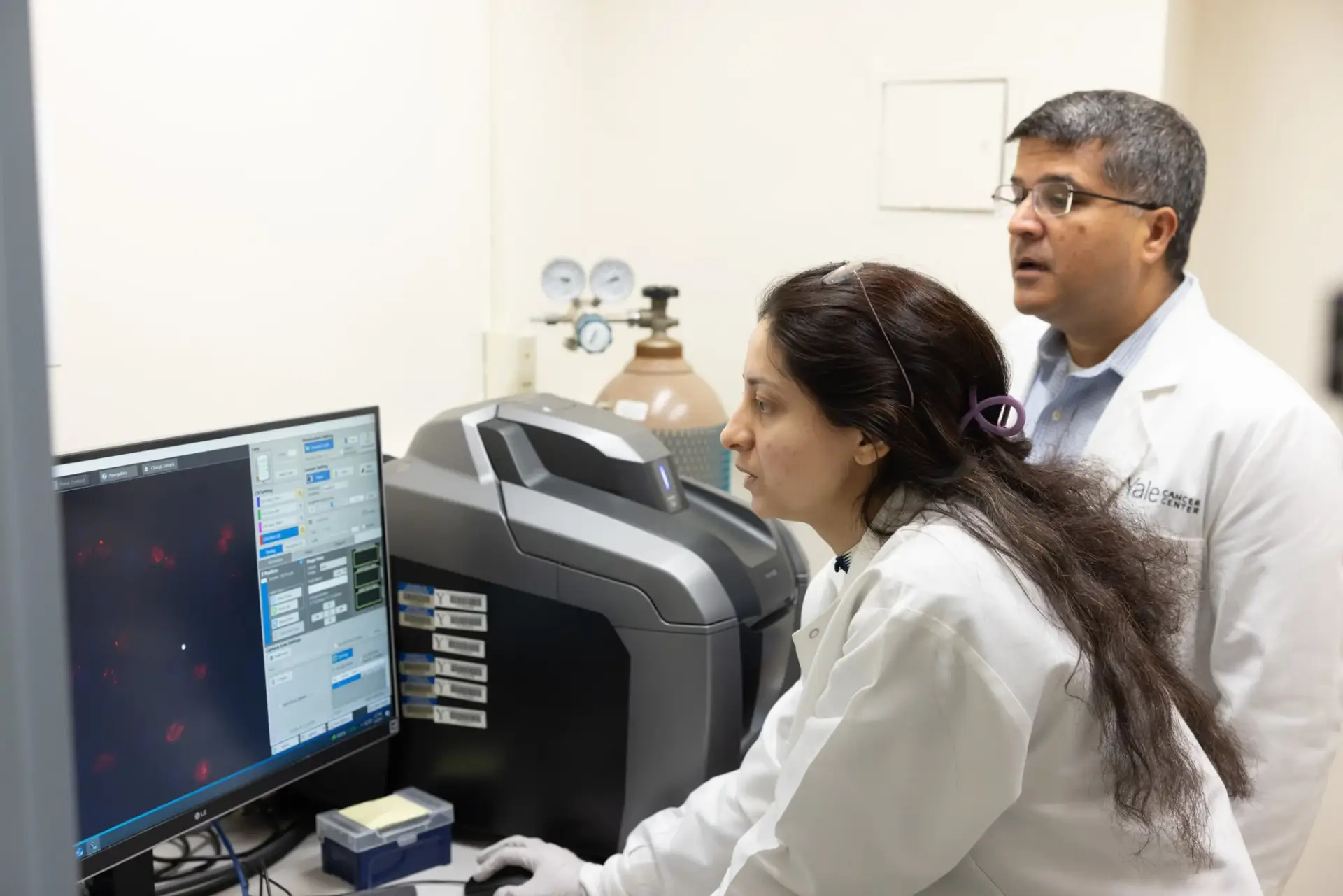 Two researchers in white lab coats review microscope images on a computer screen