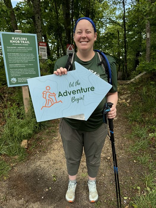 A hiker holding a Let the Adventure Begin sign on the trail